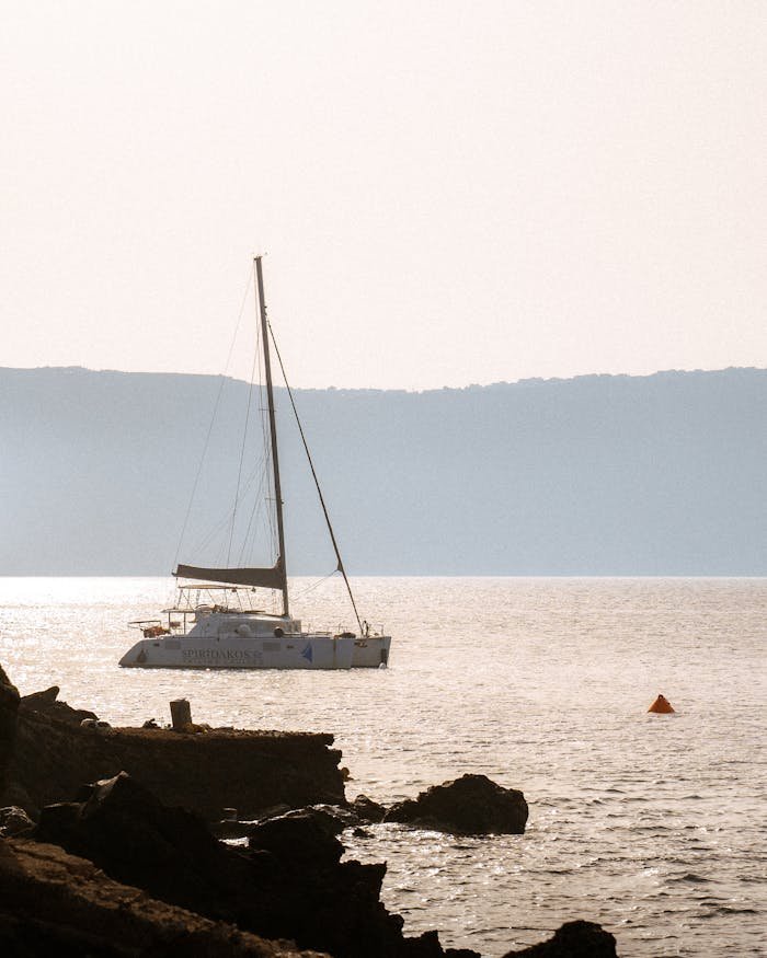 A tranquil scene featuring a yacht sailing at sunset with rocks in the foreground and a distant horizon.